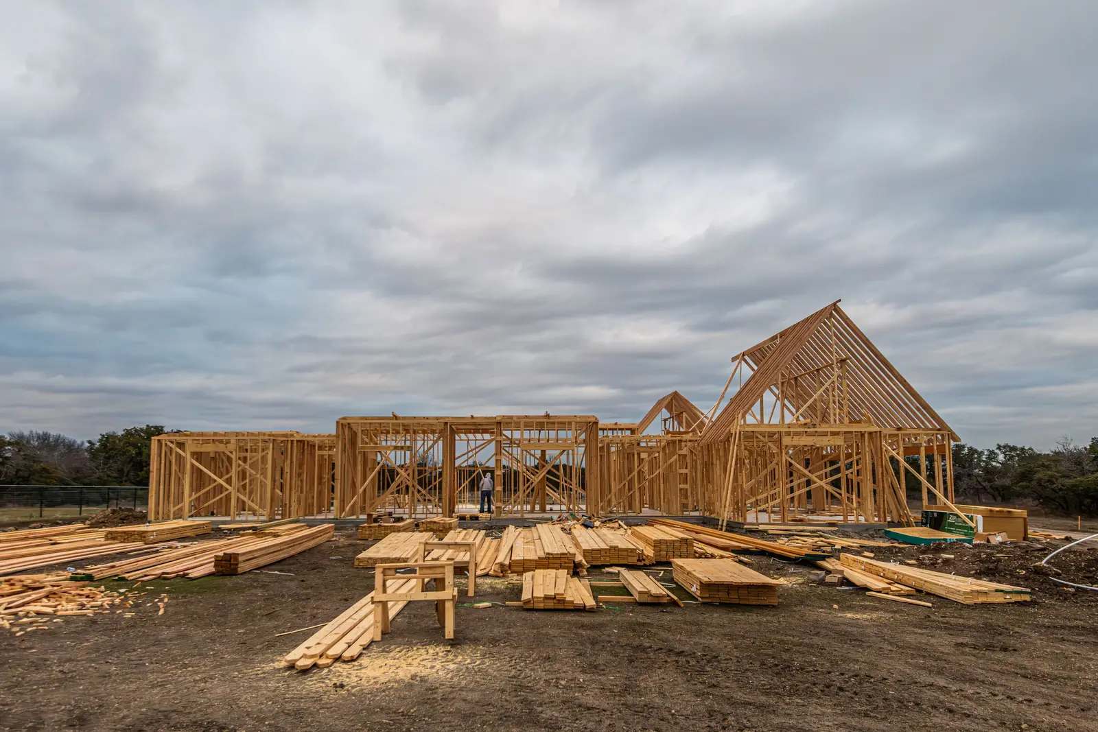 A stick-framed house with the trusses set, lumber stacked in the mud under a heavy grey sky.