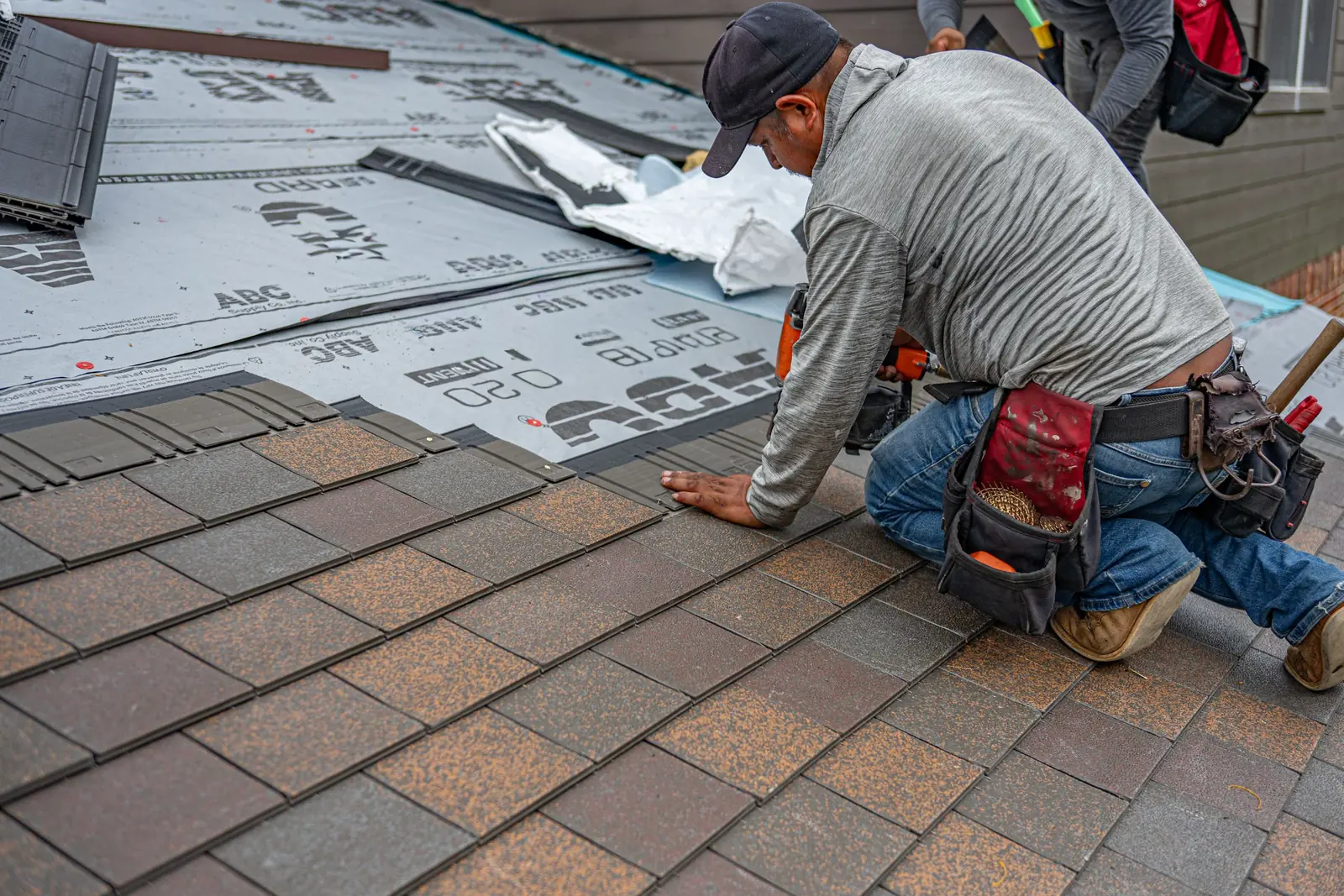 A roofer kneeling to set a course of architectural shingles over synthetic underlayment, face turned away.