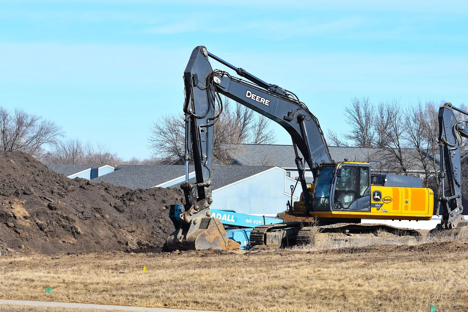 A tracked excavator working over an open trench with large-diameter pipe staged in it, spoil pile behind.