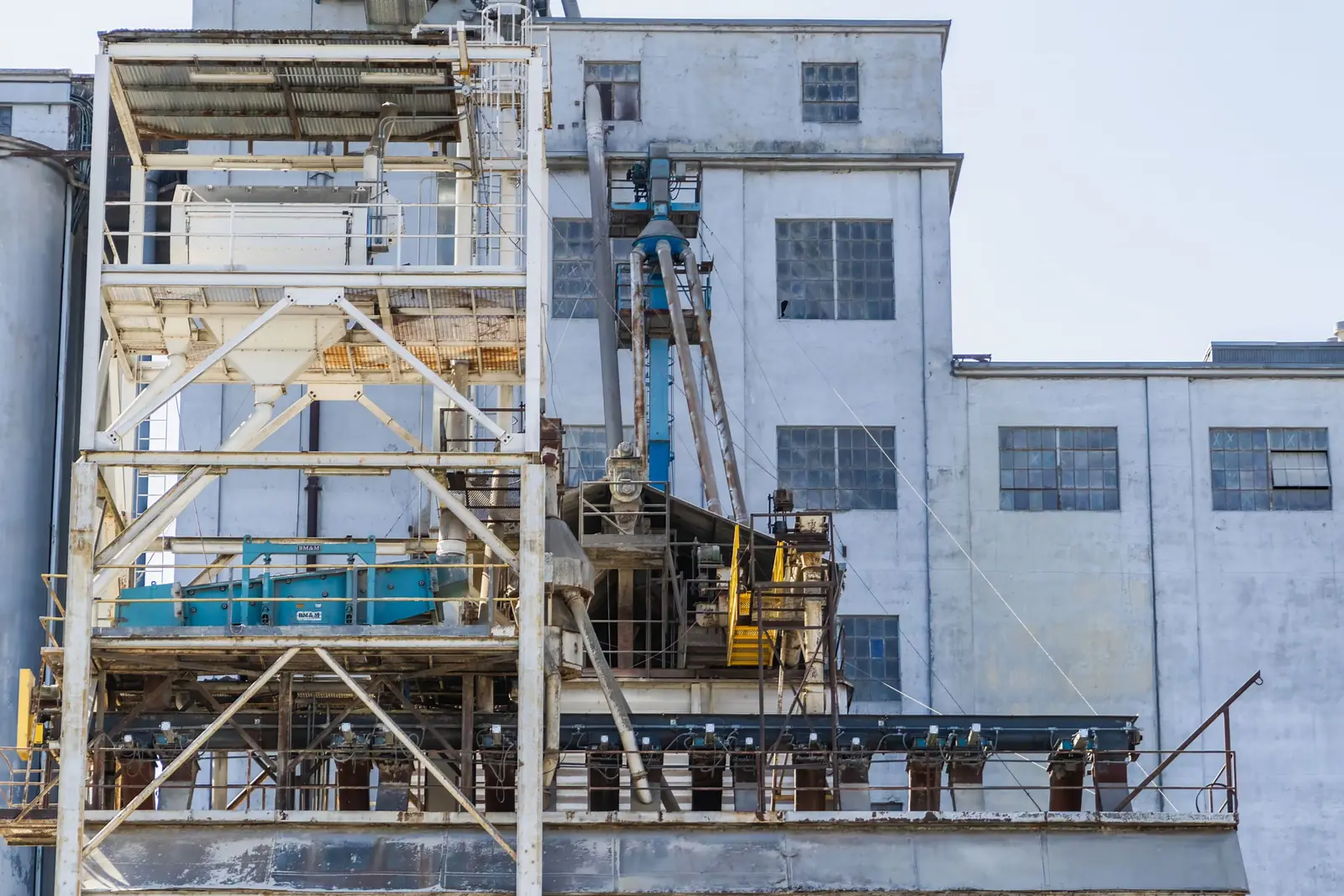 A processing plant&rsquo;s conveyor gallery, screen deck, ductwork and silos against a plain white sky.